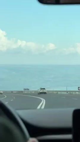 The view from inside a vehicle moving along a highway with a view of a hilly landscape and blue-tinged water. A white van is to the left, and a white Hyundai sedan, license plate partially visible, is following a white SUV with a square yellow device attached to its roof. A guardrail sits along the edge of the road. The sky is blue with white puffy clouds.