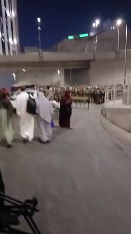 The video shows a nighttime scene on a street with many people walking. Most of the people are wearing traditional Muslim attire. A road sign indicates directions to "Abi Bakr Al Siddiq Rd." and "Jabal Al Kahba Rd.". There are motorcycles parked on the side of the street. In the background, there's a modern high-rise building with vertical stripes of light. An overpass is visible, with traffic moving above. The camera appears to be held by someone walking along with the crowd.