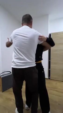 The video shows a man and a woman slow dancing in a room with white walls and light wood-look tile flooring. The man wears a white t-shirt and dark pants, and the woman wears a black hoodie, black pants with a yellow stripe, and white sneakers. A small speaker sits on the floor, and a microphone stand stands next to it. A glimpse of a mirror shows other people in the room. The camera then pans down to show a child’s foot in a pink and white sneaker. Finally, the camera focuses on a table laden with food and drinks, including chips, bread, sliced tomatoes, potatoes, and bottles of wine and beer. A phone rests on the table, displaying a video. A hand with blue nail polish hovers near the phone.