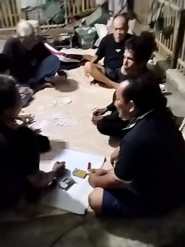 Six people are sitting on the floor in a circle playing cards. The room appears to be dimly lit and appears to be a residential or outdoor area with wooden fencing in the background. The individuals are dressed casually, with most wearing dark t-shirts and shorts. One person in the foreground is writing on a piece of paper, possibly keeping score. Cards are spread out on the mat in front of them. There are some bottles and other miscellaneous items in the background. The atmosphere seems relaxed and social.
