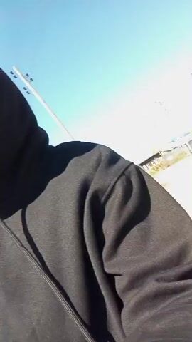 A woman with reddish-brown hair is featured in a close-up, selfie-style video. She is wearing a black zippered hooded jacket with the hood up, and large, dark rectangular sunglasses. A white embroidered logo or crest is visible on the left chest area of her jacket, though the text is illegible. She has subtle makeup and appears to be smiling faintly. In the background, a clear, bright blue sky is prominent, suggesting a sunny daytime setting. In the initial frames, a blurry background behind her shows a light-colored fence and some distant, light-colored structures, indicative of an outdoor, possibly semi-rural or industrial environment in Yurgamysh, Russia. The woman is looking directly at the camera, and the video primarily captures her upper body and face as she slightly shifts her head and the camera angle.