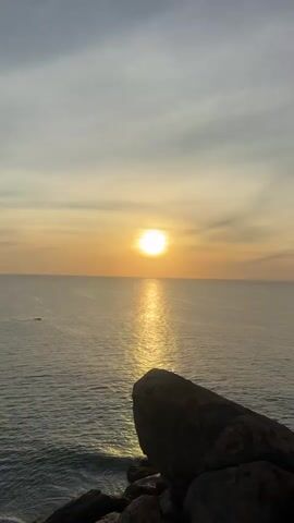 A static, vertical shot captures a serene ocean sunset. The sun, a bright, white orb, hangs low in the sky, casting a shimmering golden reflection across the calm water. Wispy clouds, tinged with orange and yellow, fill the sky, suggesting a peaceful end to the day.

In the foreground, a large, dark rock formation juts out into the frame from the bottom right, providing a natural frame for the expansive seascape. The rock appears weathered and solid, anchoring the scene against the vastness of the ocean and sky.

The water is a deep, tranquil blue, with gentle ripples visible, reflecting the soft light of the sunset. A small, dark object, possibly a boat or a buoy, is faintly visible in the distance on the left side of the frame. To the far left, a faint landmass can be seen on the horizon.

The overall mood is one of tranquility and natural beauty. The time of day is clearly sunset, given the position of the sun and the warm hues in the sky. The location appears to be a coastal area with rocky outcroppings, consistent with Shastrawela, Sri Lanka. There is no visible text in the image.