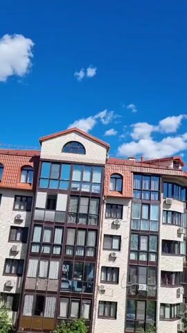 This vertical video displays a view from a window overlooking an urban or suburban environment. The primary subject is a multi-story building with a distinctive curved facade and a terracotta-tiled roof. The building's exterior is a light-colored material with numerous large windows, many of which are reflective, showing the blue sky and clouds. Air conditioning units are visible mounted on the exterior walls.

Below the building, a street or parking area is visible, with several cars parked. These include a white SUV, a dark sedan, a white sedan, and a yellow car. A person can be seen walking near the cars in the distance. Green trees and foliage surround the area, with a line of trees forming a backdrop to the buildings and cars. In the far distance, more buildings and structures are visible, suggesting a populated area.

The sky is a vibrant blue with scattered white clouds, indicating fair weather. The sunlight suggests it is daytime, likely late morning or afternoon due to the brightness and shadows. The overall impression is a peaceful, everyday scene, possibly from a residential building. No text is discernible in the video. The location context is Romashkovo, Russia.