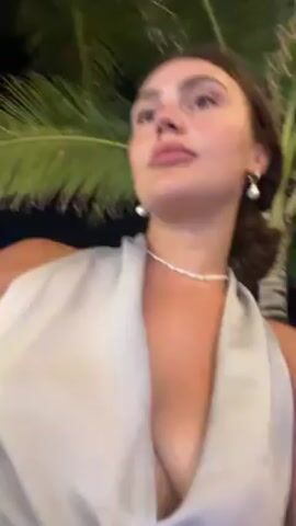 A woman is outdoors at night with palm trees visible. She has her hair in a low bun and is wearing pearl earrings and a pearl choker, along with a light grey or silver dress. She takes a drink from a dark brown bottle with gold labeling.