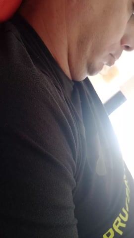 A low-angle, close-up shot features the profile of a man wearing a black t-shirt. The man's face is partially visible, showing the bottom half including his lips, chin, and part of his nose. He appears to be looking down, possibly at something in front of him. The background is bright, with a rectangular window visible. The man is positioned on a red surface, possibly a chair or couch. The t-shirt has a word printed along the sleeve, though the full word is cut off, with only "PRUEBA" visible.