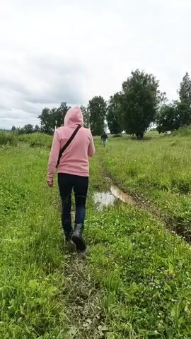 The video shows two people walking along a muddy path through a grassy field. The person in the foreground is wearing a pink hoodie and black pants, while the person in the background is wearing a blue shirt. There are trees and a water tower in the distance. The sky is cloudy. The video appears to have been filmed in daylight.