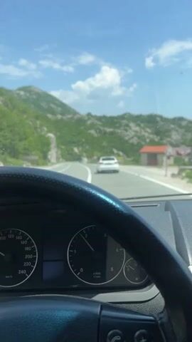 The video shows the dashboard and steering wheel of a car driving on a road, with a white car in front. The road is lined with trees and bushes, and there are mountains in the background. The sky is blue with white clouds. The car is driving at a speed of 70 km/h. The road has white lines marking the lanes. There are road signs indicating a curve ahead and a speed limit.