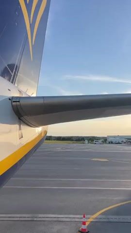 The video shows a close-up view of the tail and wing of a Ryanair airplane from an external perspective. The plane is on an airport tarmac. In the background, a control tower and several airport buildings are visible under a blue sky with scattered clouds, bathed in the warm light of what appears to be either sunrise or sunset. The camera pans slightly to the right, revealing more of the tarmac and the airport infrastructure. There are no people visible in the video.