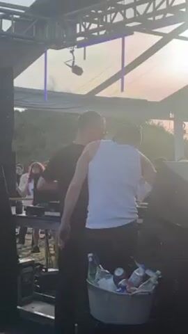 The video shows a DJ set in a partially enclosed area with a roof structure made of metal beams and translucent panels. The light suggests either dawn or dusk. The crowd of people is visible in front of the DJ booth, dancing and enjoying the music. The booth is occupied by two men. One is wearing a white tank top and black pants and is standing behind the other. The other man is wearing a black t-shirt and is operating the DJ equipment. There is a bucket filled with ice and drinks in front of them. The area is smoky, possibly from a fog machine, with blue LED lights hanging from the roof structure. The atmosphere appears to be festive and energetic.