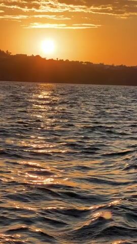 The video shows a sunset over water. The sun is a bright orb in the center of the frame, casting a golden glow across the water's surface. The sky is orange, with some wispy clouds near the horizon. The water is dark and choppy, with small waves reflecting the sunlight. There is a silhouette of land visible in the distance. As the video progresses, the camera moves, creating splashes of water on the side.