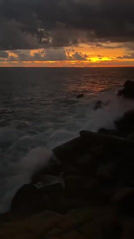 This media file captures a dramatic coastal scene at sunset or sunrise. The horizon is illuminated by a vibrant band of orange and yellow light, indicating the sun is very low. Above this, the sky is filled with heavy, dark grey clouds, creating a striking contrast between the intense light and the brooding atmosphere.

In the foreground, large, dark rocks form the coastline, extending from the right side of the frame. Powerful waves crash against these rocks, generating significant white foam that stands out against the dark water. The ocean itself appears dark and deep, reflecting some of the warm hues from the sky.

The scene conveys a sense of natural power and beauty, with the turbulent sea meeting the solid rock under a dramatically lit sky. No people, visible text, or man-made structures are present, focusing entirely on the raw elements of nature. The setting is a rocky coast, consistent with the provided location of Rumassala, Sri Lanka.
