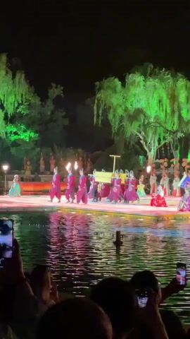 A nighttime shot shows a performance happening over a body of water. Performers dressed in matching red and blue costumes carry a covered object on poles across the stage. The stage, set over the water, features trees lit in green and decorative structures in the background. The audience is visible in the foreground, many recording the performance on their phones. The water reflects the lights and stage elements, adding to the spectacle.