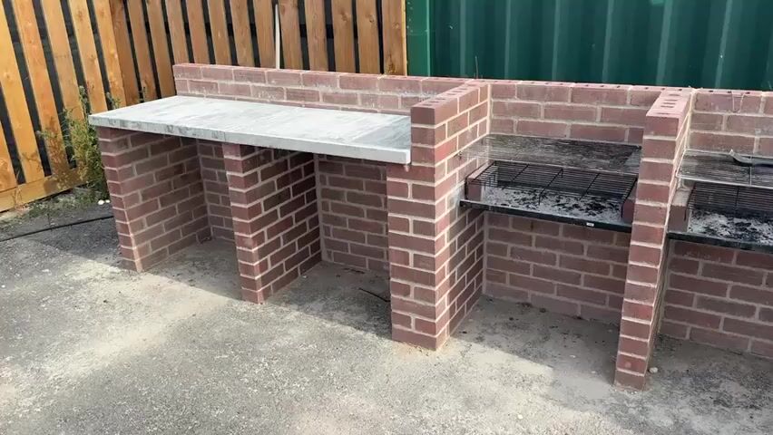 The video shows an outdoor scene. In the foreground, there is a brick structure that appears to be a built-in barbecue or outdoor kitchen. It has three main sections. The leftmost section has a flat, light-colored countertop. The two sections to its right have metal grates set into them, suggesting they are for cooking, and there is evidence of ash or charcoal on the grates. The barbecue structure is made of red bricks and has openings beneath the cooking areas and countertop. The ground around the structure is paved with concrete or gravel.

In the background, there is a large green shipping container with the logo "MBOX" and a phone number "0800 783 9885" visible on its side. To the right of the shipping container, there are stacks of building materials, including insulation boards and a yellow container. Further in the background, there is a small wooden shed, some fencing, and a light-colored cylindrical structure, possibly a silo. The overall environment appears to be an outdoor construction or storage area, with some vegetation visible. The lighting suggests it is daytime, possibly a sunny day. There are no people visible in the video. The camera pans slowly from left to right, showing the barbecue structure and its surroundings.