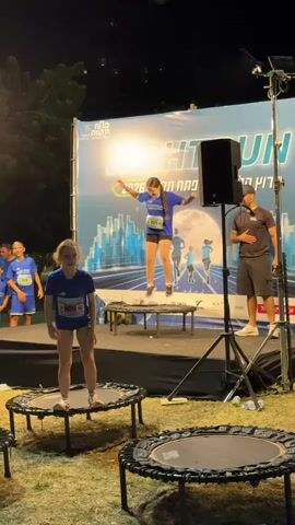 This video captures an outdoor night event in Petah Tikva, Israel, featuring a trampoline jumping activity. The scene is illuminated by artificial lights, highlighting a stage with a large backdrop banner and several small trampolines set up on a grassy area.

On the stage, a young girl in a blue t-shirt and shorts is energetically jumping on a trampoline, reaching considerable height. To her right, a man in a black t-shirt and shorts, seemingly an instructor or event staff, stands and gestures, observing the activity. A large black speaker on a stand is positioned next to him. In the foreground, another young girl wearing a blue t-shirt with the number "027" and shorts stands on a smaller, circular trampoline placed directly on the grass. Other children in similar blue attire are visible near the stage, watching the proceedings.

The atmosphere appears vibrant and engaging, with participants actively involved in the trampoline fun.

The prominent banner on the stage displays "NIGHT RUN" in large, stylized letters. Below this, text in Hebrew reads "פתח תקווה 2026" (Petah Tikva 2026) and "רץ עם תקווה" (Running with Hope). The banner's design includes a graphic of runners silhouetted against a city skyline with a large full moon, reinforcing the "Night Run" theme. The event takes place at night, as indicated by the dark sky and the reliance on stage lighting. The children's blue shirts also feature visible logos and numbers, typical of participants in an organized race or festival.