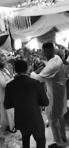 A black and white video captures an indoor social event with multiple attendees dancing. In the foreground, a woman wearing a dark, patterned traditional dress with elaborate beading around the neckline dances in the center. To her immediate right, a man in a light-colored traditional outfit holds a microphone to his mouth, appearing to sing or speak, while facing and engaging with the dancing woman. To the left of the central woman, another woman in a light-colored traditional outfit is seen dancing and smiling. Numerous other individuals are visible in the mid-ground and background, also participating in the dancing or observing the scene. The venue features draped fabrics and long, decorative strings or beads hanging from the ceiling, indicating an ornate event setting.