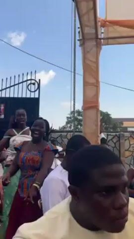 The video shows a group of people dancing outside on a sunny day. Most of the dancers are women wearing colorful African-style dresses. One woman is wearing a green dress, another a pink top, and one has on a blue and orange print dress with a maroon skirt.  Some of the dancers are smiling and laughing. There is a wrought iron gate in the background, as well as a draped pillar that may be part of an event setup. A few men are present but not actively dancing.  A small child with curly hair is visible, also appearing to enjoy the music.