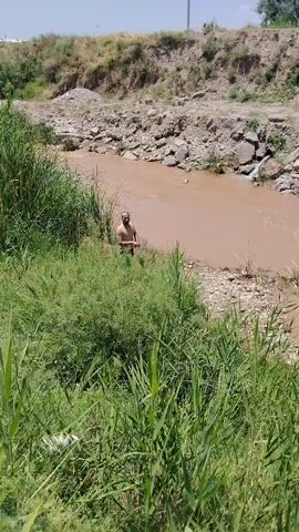 A man stands in shallow, murky brown water near the edge of a riverbank. He is wearing dark shorts. He walks out of the water onto a rocky shore. Another man, also wearing dark shorts, walks into the river from the shore. The riverbank is covered with dry, brown soil and rocks. Green reeds and tall grass grow along the left bank of the river.