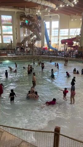 This video shows an indoor water park with a large wave pool. Many people, including adults and children, are playing in the water, some are standing, others are sitting or lying down. There are several water slides visible in the background, and a staircase leading up to them. The environment appears to be well-lit with natural light coming from large windows and artificial lighting. The water is clear and there are waves present in the pool.