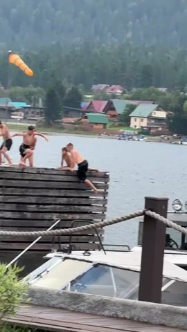 Four young males are on a wooden pier. The pier is elevated above the water, with a boat docked alongside it. In the background, there are trees, houses, and a body of water. The sky is overcast. One male is standing on the left side of the pier, facing forward. Two males are crouched down in the middle of the pier, one leaning on the other. Another male is sitting on the edge of the pier on the right side, facing the water. There is a windsock on a pole on the left side of the pier.