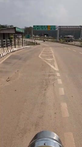 The video shows a POV shot from a vehicle, likely a motorcycle, traveling on a paved road. The road is marked with white chevron markings pointing forward. On the left side of the road, there is a sidewalk with a metal railing and a green sign with white text. Further down the road, there are multiple traffic signs. One large green overhead sign has directional information with city names like Dharamshala, Kangra, Mandi, and Kotla, along with their distances. The sign also indicates NH 134. In the background, there are trees and hills under a cloudy sky. A few vehicles, including a bus, are visible in the distance. The general environment appears to be an approach to a highway or a major intersection.