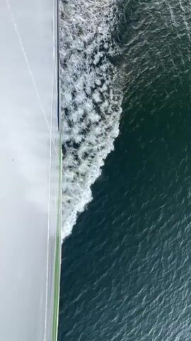 This is an aerial, high-angle shot of the side of a large ship moving through dark blue water. The ship's hull is white with a thin green stripe near the waterline. The water is disturbed by the ship's movement, creating a white frothy wake that trails behind. The wake is widest near the ship and tapers off as it moves away. The water itself appears choppy with small waves and ripples. The lighting suggests it is daytime, possibly late afternoon or early morning, given the warm, golden hue reflecting off the water in the distance. The scene is static, with no people or other objects visible. The camera appears to be stationary and looking directly down at the water and the ship's side.