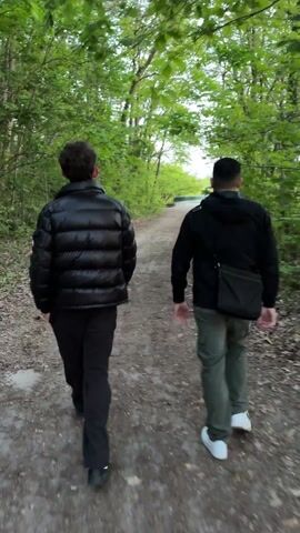 The video shows two men walking away from the camera on a dirt path through a wooded area. The man on the left wears a black puffer jacket and black pants, and he has glasses. The man on the right wears a black jacket, green pants, and white shoes, and has a black shoulder bag. The path leads towards a clearing with a glimpse of water in the distance. The surrounding trees have green leaves.