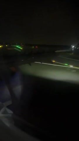 The video shows a view from an airplane window at night. The runway lights are visible, and another airplane is seen on the runway in the distance. The wing of the airplane the video is being filmed from is also visible. The scene is dark, with the lights of the runway and the other airplane being the primary sources of illumination.