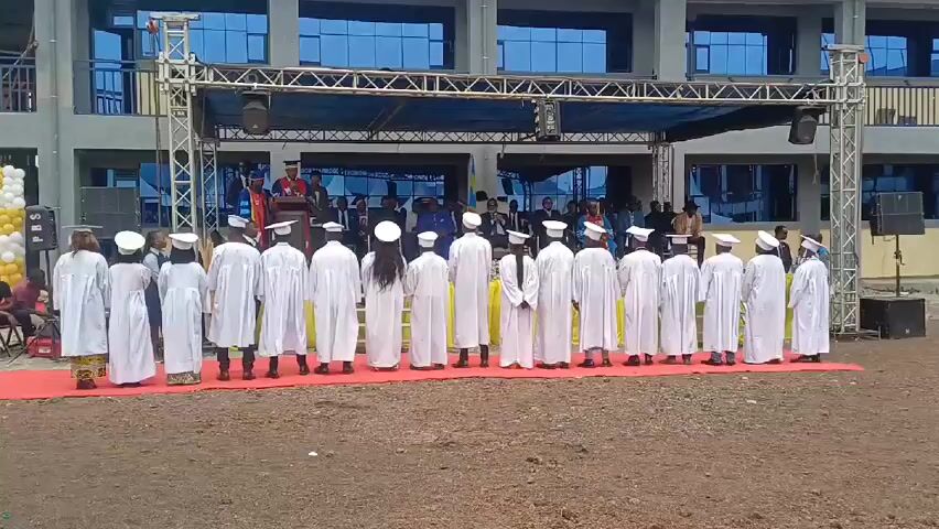 The video shows a graduation ceremony taking place outside in front of a building.  A group of students in white robes and caps are standing in a line on a red carpet.  They are facing a stage where people are speaking.  There are balloons to the left.  The building behind the stage has many windows.