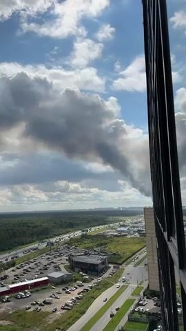 The video shows a wide shot of a landscape with a tall building in the foreground on the right. A plume of dark smoke is visible in the sky, rising from the horizon and spreading across a patch of clouds. Below the smoke, a dense forest stretches out, with a highway running through it. Cars and trucks are seen driving on the highway. In the mid-ground, there are several buildings, parking lots filled with cars, and smaller roads. The sky is blue with scattered white and gray clouds. The camera appears to be positioned high up, possibly from a window or balcony of the building.
