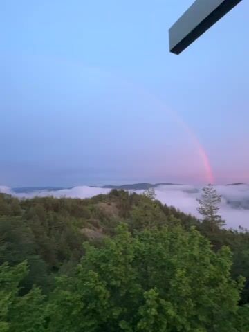 A video shows a landscape scene with a rainbow arching across the sky above a valley filled with clouds. The foreground is dominated by lush green trees. In the upper right corner of the frame, a portion of a metallic structure is visible, possibly part of a viewing platform or a cross. The sky transitions from a pale blue to hues of pink and orange, suggesting either dawn or dusk. The overall impression is serene and natural.
