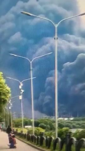 A plume of thick, dark smoke billows into a pale sky, obscuring a distant landscape of green trees and industrial structures with tall, slender towers. In the foreground, the tops of two streetlights with modern, elongated fixtures are visible, one positioned centrally and the other to the left. A low barrier, possibly a fence or railing, runs along the bottom edge of the frame. The smoke is the dominant element, suggesting a large fire or industrial event.