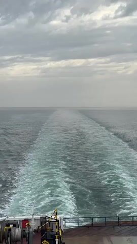 The video shows the view from the back of a moving vessel, likely a ship or ferry, out over the water. The sky is overcast with gray clouds. The water is mostly dark with a lighter, foamy wake stretching far into the distance behind the vessel. Some machinery, including a winch and railing, is visible on the deck of the vessel in the foreground.