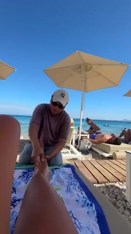 A person is getting a foot massage on a beach. The masseuse, wearing a purple shirt and a white baseball cap, is kneeling and massaging the person's foot. The person receiving the massage is lying on a beach chair, covered by a blue and white towel with a coral pattern. In the background, there are other people on lounge chairs under umbrellas, and the ocean with clear blue water and a bright sky.