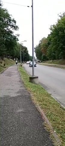 A video shows a paved sidewalk next to a two-lane road. The sidewalk is on the left side of the frame, and the road is on the right. A lamppost is visible in the foreground on the right side of the sidewalk. Trees line both sides of the road. A person is walking on the sidewalk in the distance. Several cars are driving on the road, including a black SUV, a silver sedan, and a white van. The sky is overcast.