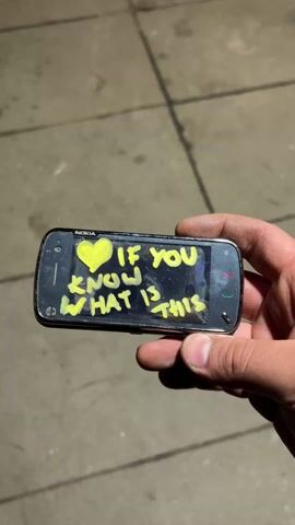 A person is holding a Nokia N97 mobile phone. The screen has yellow graffiti, a heart, and the message, "If you know what is this". The phone has a slide-out keyboard that is being exposed. The phone is held over a concrete ground.