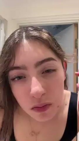 A medium close-up shot shows a young woman looking into the camera as she uses an eyelash curler on her right eye. She has fair skin, brown hair with bangs, and wears a black tank top. She has a nose ring and a necklace with a circular pendant. Her right hand holds the eyelash curler, and her fingernails are painted with pink and silver polish. The bathroom door is visible in the background.