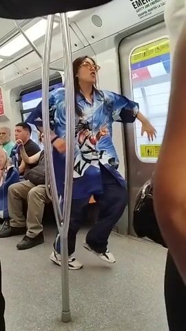 Inside a train car, a woman with glasses and a blue shirt with anime characters is dancing. She moves her arms and head, swaying to the music. Several passengers are sitting on the train seats, some looking at her. A crutch is leaning against a support pole. Other passengers are standing near the camera, partially blocking the view. The train car has advertisements and safety notices.