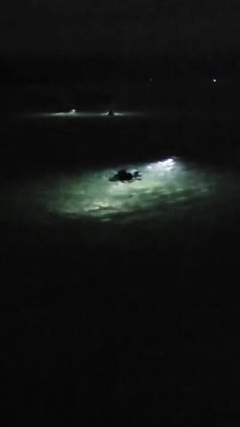 At night, a figure swims in water illuminated by a bright light. In the background, more lights suggest other figures or vessels, moving horizontally across the dark water. The scene is dark, and the primary focus is on the illuminated swimmer.