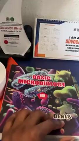 A hand flips through the pages of a textbook titled “Basic Microbiology for Students” while on top of a blue desk. In the background are an Award of Merit trophy and a 2024 calendar.
