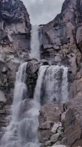 A waterfall cascades down a rocky cliff face. The water falls in multiple streams, some wide and powerful, others narrow and delicate, over large boulders and jagged rock formations. The surrounding cliffs are steep and rugged, with visible layers of stone and patches of green vegetation clinging to the rock. The sky above is overcast with grey clouds. The camera appears to be positioned at the base of the waterfall, looking upwards at the impressive natural spectacle.