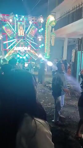 The video shows a crowd of people gathered in what appears to be an outdoor event at night. There is a large, elaborate light display, possibly a stage or a sound system, with bright, colorful lights flashing. People are standing and some appear to be dancing or moving to music. The lighting is dynamic and intense, with beams of light cutting through the darkness. The ground is uneven and appears to be dirt or gravel. The overall atmosphere suggests a lively celebration or performance.