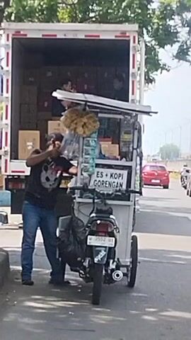 A man in a black t-shirt and blue jeans stands next to a mobile food cart attached to a motorcycle. The cart displays a sign that reads "ES + KOPI GORENGAN". He is holding a cup and appears to be interacting with the cart. Behind the cart, a white truck is parked with its back doors open, revealing boxes stacked inside. Two individuals are visible inside the truck. In the background, several motorcycles are seen on a road, with some vehicles also present. The environment appears to be outdoors, possibly near a street or market.