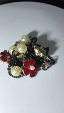 The media file is a close-up video of a delicate piece of jewelry. The object is a necklace, featuring a dark, intricate chain interspersed with several pearl-like beads and small, red enamel flower charms. Each flower charm has a tiny white bead at its center, resembling a stamen. The metal components of the necklace appear to be a dark, possibly antique brass or pewter tone, with gold-toned findings for the clasp and links. The pearls are lustrous and have a creamy white hue. The necklace is arranged in a loose pile on a clean, white surface, which is likely a table or display stand. The lighting is soft, creating gentle shadows that highlight the texture and form of the jewelry. There is no visible text in the media file. The scene is static, with the necklace simply resting in place, suggesting it is being presented or photographed. The overall impression is one of elegance and classic design.