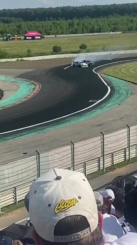 The video shows a motorsports event from the perspective of spectators. In the foreground, a person wearing a white baseball cap is visible, partially obscuring the view. Beyond the spectators, a racetrack is seen with several race cars drifting, producing large amounts of smoke. The track is bordered by green and black asphalt with white lines. In the background, there are trees, grassy fields, tents, and vehicles. The sky is bright and appears to be daytime with some clouds. The camera is held at an angle, suggesting it is being used to record the event from a spectator's viewpoint.