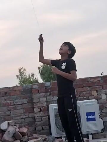 A young boy is standing outdoors on a brick wall structure, possibly a rooftop or terrace. He is wearing a black t-shirt and black pants. The boy is holding a string that extends upward and out of frame, suggesting he is flying a kite. He looks up and gestures with his hands as if guiding the kite. Behind him is a brick wall and a Haier air conditioning unit. In the background, there are trees and a cloudy sky.