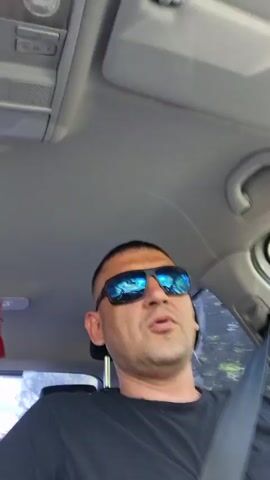 A man wearing sunglasses and a black t-shirt is sitting in the driver's seat of a car. He is looking at the camera and speaking. The interior of the car is visible, including the ceiling and the dashboard. Outside the car, trees and foliage can be seen. The man appears to be in motion, suggesting the car is being driven.