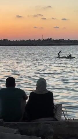 Two people sit facing away from the camera on the shore of a body of water. The person on the left wears a dark green shirt. The person on the right wears a black jacket and a light-colored hijab. Next to the person on the right is a woven tote bag. The water has small waves. In the distance, two people are on a kayak or small boat. One person stands and holds a paddle, and the other person is seated. A rocky structure extends into the water behind the boat. The sky has a sunset gradient with orange and pink hues and scattered clouds.