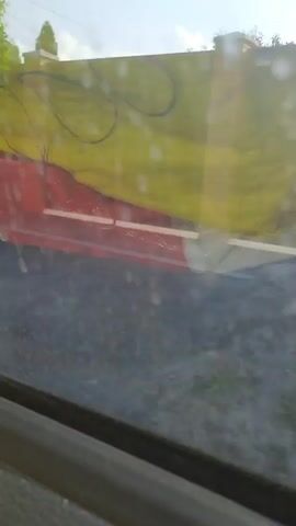 This is a video taken from inside a vehicle, likely a car or bus, looking out a window. The scene is viewed through a dirty window, which distorts the image and adds a hazy, speckled effect.

The primary subject is a landscape that appears to be in a rural or semi-rural setting in Sadky, Ukraine. In the foreground, through the window, we see the interior of the vehicle, including a dark grey window frame and a textured surface that might be a car seat or dashboard.

Outside the window, the dominant visual elements are a large, horizontal mural or painted wall. This mural features bold, horizontal stripes of color: red on the bottom, followed by yellow or light green, and then a darker green or blue at the top. The mural appears to be on a fence or wall that stretches across the frame. Behind the mural, we can see trees with green foliage, suggesting it is daytime and likely spring or summer. The sky is visible in the upper portion of the frame and appears bright, indicating it's likely daytime. There are also some structures visible, including what might be houses with roofs and possibly a utility pole. In some parts of the video, there are mounds of dirt or sand in front of the mural, suggesting ongoing construction or landscaping.

The activity taking place is simply the vehicle moving through this location. The perspective of the video suggests the viewer is a passenger. There are no people clearly visible, and the focus is on the scenery passing by.

Notable details include the dirty window, which significantly impacts the clarity of the scene. The colors of the mural are vibrant despite the distortion. The lighting suggests a sunny day. The location cues confirm it is Sadky, Ukraine. No text is discernible in the video. The overall mood is observational, capturing a fleeting glimpse of the environment.