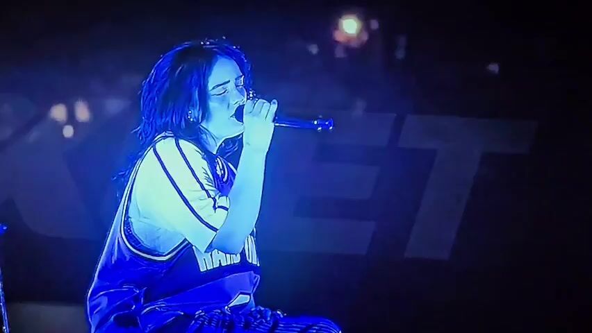 A medium shot shows a young woman with dark, shoulder-length hair and closed eyes, holding a microphone to her mouth with her right hand and singing. She is wearing a white t-shirt under a blue basketball jersey with white trim and lettering. She is also wearing striped pants. The scene is lit with a strong blue light, creating a silhouette effect. In the background, a large, blurry, white logo or text is visible, partially obscured by the blue light. The background appears to be a dark, out-of-focus concert venue with some distant lights.