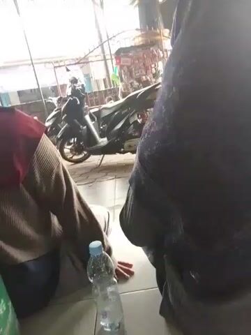 The video shows a person sitting on the floor, with a plastic water bottle in front of them. Another person, wearing a dark hijab, is sitting to the right, obscuring most of their body. In the background, a motorcycle is parked. The environment appears to be an outdoor seating area, possibly a market or food stall, with tiled flooring and a covered roof structure overhead. The lighting is bright, suggesting daytime.